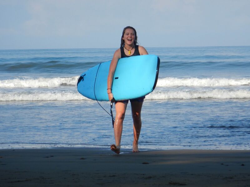 A woman is walking on a beach towards the camera, carrying a blue surfboard under her arm. She is smiling and appears to be enjoying herself. The ocean is behind her with small waves breaking on the shore. The sky is bright and clear, suggesting a sunny day. The sand is dark and wet, indicating the tide is low. She is wearing a dark swimsuit and flip-flops.